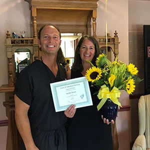 Dr. John Louis standing alongside a female patient who is holding a framed Patient of the Month certificate and an arrangement of sunflowers.