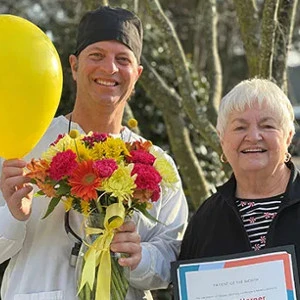 Dr. John Louis presenting a smiling female patient with a certificate, yellow balloon, and a colorful floral bouquet.