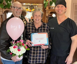 A dental staff member and doctor presenting a female patient with a Patient of the Month certificate, a pink balloon, and a bouquet of flowers inside the office.