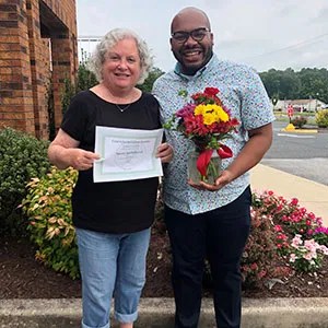 A male standing outdoors with a female patient holding her certificate alongside a vase of fresh flowers.