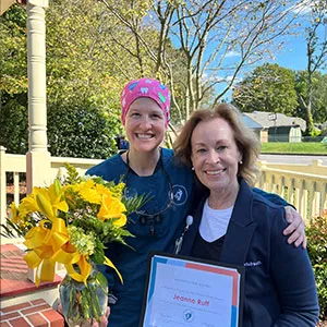A female staff member in scrubs standing outdoors with a patient who is holding a certificate and a bouquet of yellow flowers.