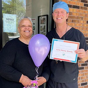 Dr. John Louis standing next to a smiling female patient who is holding a purple balloon and a framed certificate.