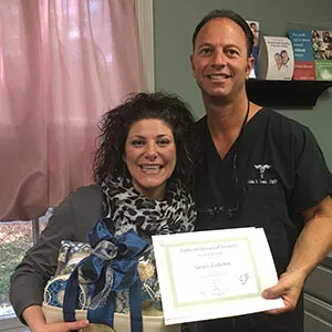 Dr. John Louis standing with a smiling female patient who is holding a gift basket and a Patient of the Month certificate.