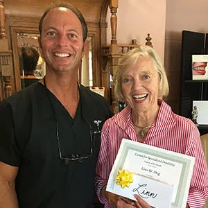 Dr. John Louis smiling alongside a female patient wearing a pink collared shirt, who is proudly holding her framed certificate.