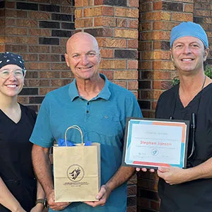 A male patient holding a gift bag, posing alongside Dr. John Louis holding a certificate and a female staff member in front of a brick building.