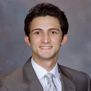 Studio portrait of Dr. Georgi Talmazov smiling, wearing a suit jacket, white dress shirt, and patterned tie against a gray backdrop.