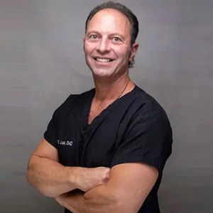 Studio portrait of Dr. John Louis smiling with arms crossed, wearing black medical scrubs against a solid gray backdrop.